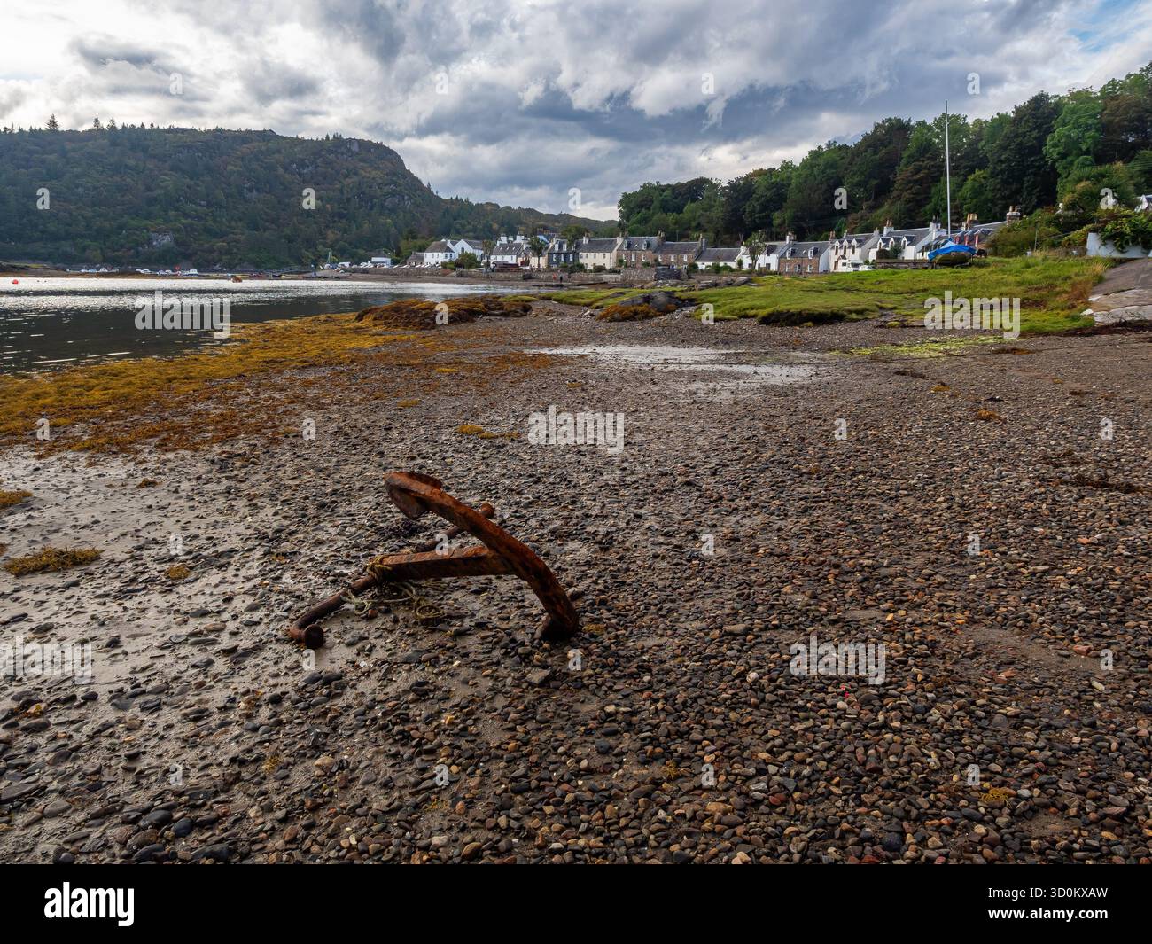 Rusty anchor on pebbled foreshore with Plockton village and hills beyond Stock Photo