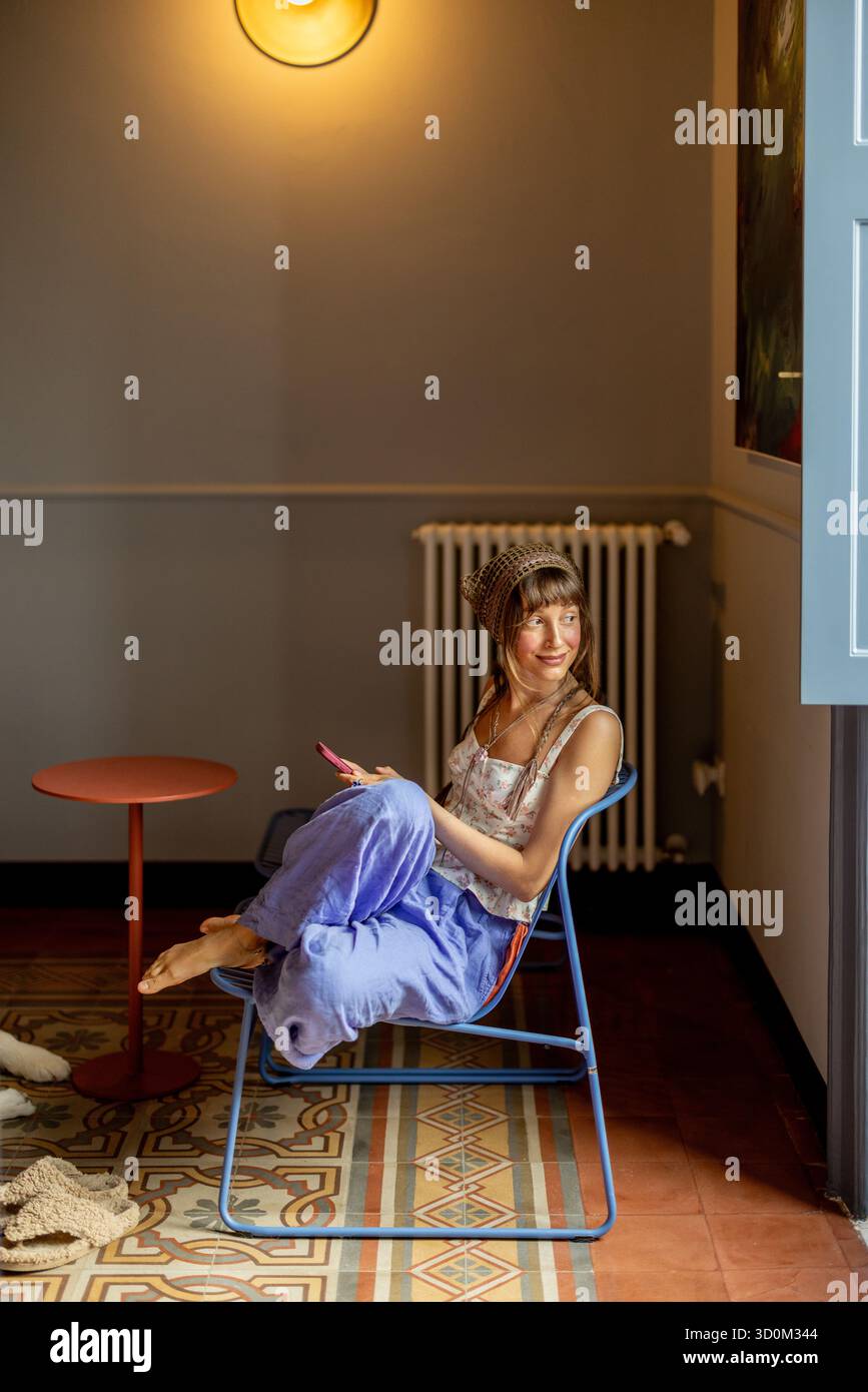 Woman sitting barefoot on a chair in a cozy Sicilian apartment, surrounded by warm light and vintage tiles. Peaceful slow living moment in Italy Stock Photo