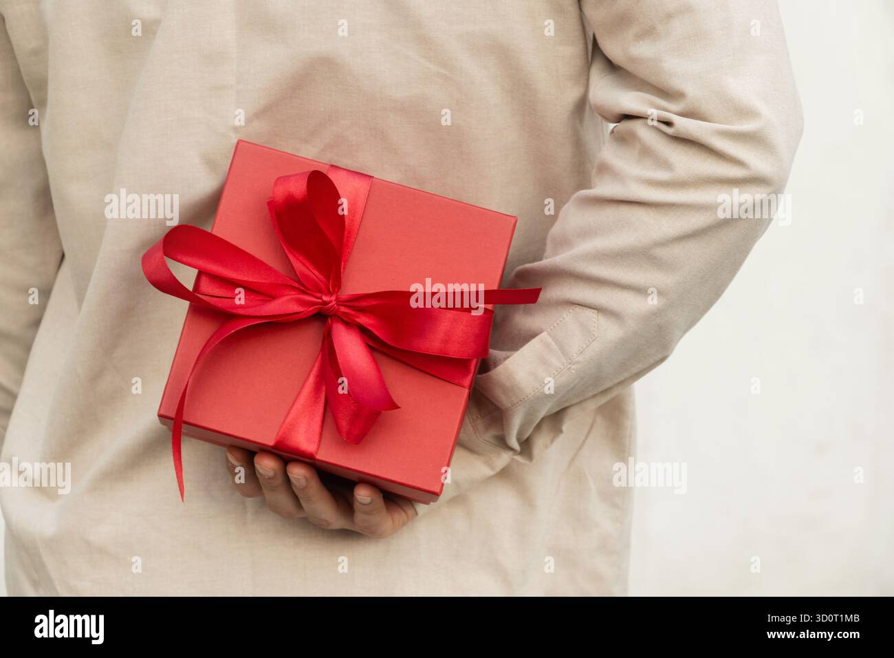 Man hiding red gift box tied with ribbon behind their back, wearing beige shirt for surprise girlfriend. Birthday present suggests surprise or special Stock Photo
