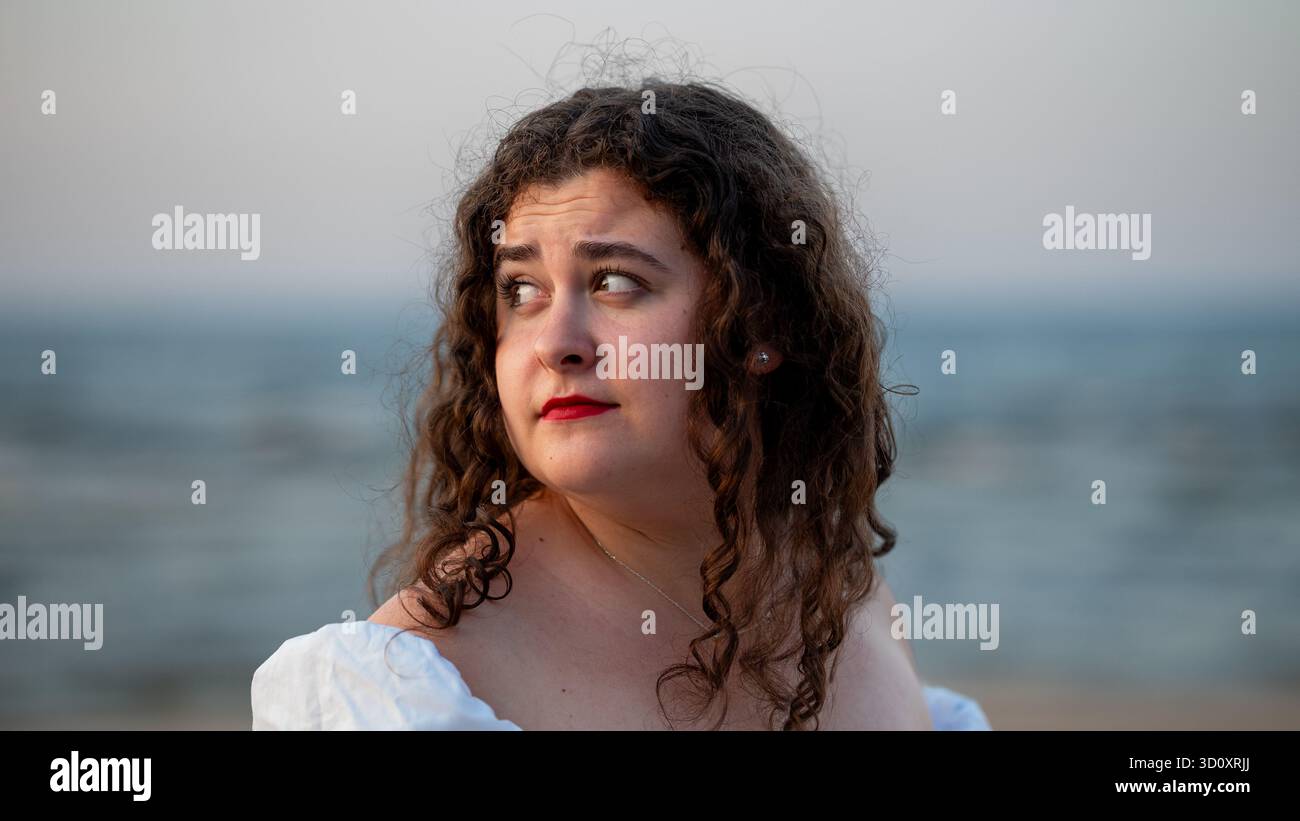 Young woman with curly hair gazes thoughtfully at the ocean during a serene beach sunset Stock Photo