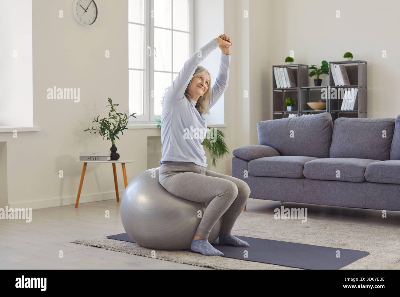 Old grey haired woman exercising with fitball, enjoying gymnastics at home, keeps arms above head Stock Photo
