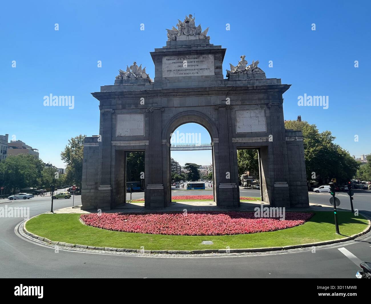 Front view of the Puerta de Alcalá in Madrid, Spain, surrounded by pink summer flowers on a sunny day. - Smartphone Captured Stock Image