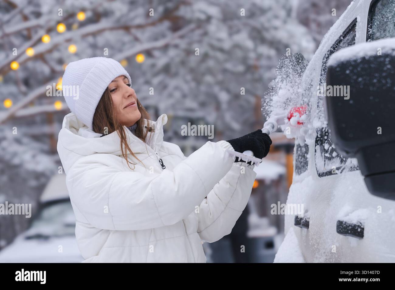 A 30- to 35-year-old woman in a white jacket and hat joyfully and with a smile on her face cleans the stuck snow and ice from her car, getting ready Stock Photo