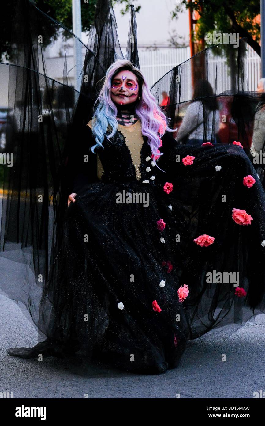 Woman in elaborate black dress with floral accents and colorful hair wearing Day of the Dead face paint during street celebration Stock Photo