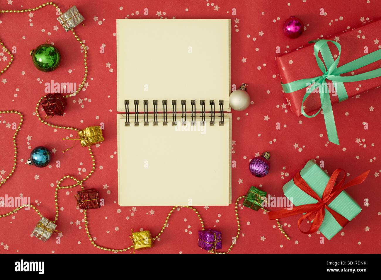 Open blank notebook in the center surrounded by Christmas tree branches, holiday decorations, and gifts on a red background Stock Photo