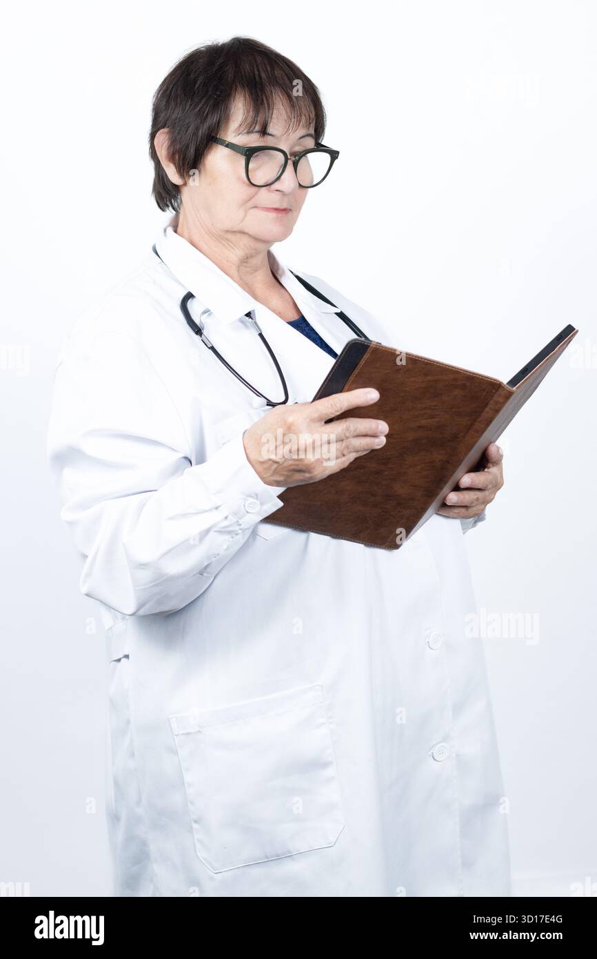 Mature female doctor in lab coat and stethoscope, wearing glasses, standing and studying a patient record book—focused, professional Stock Photo