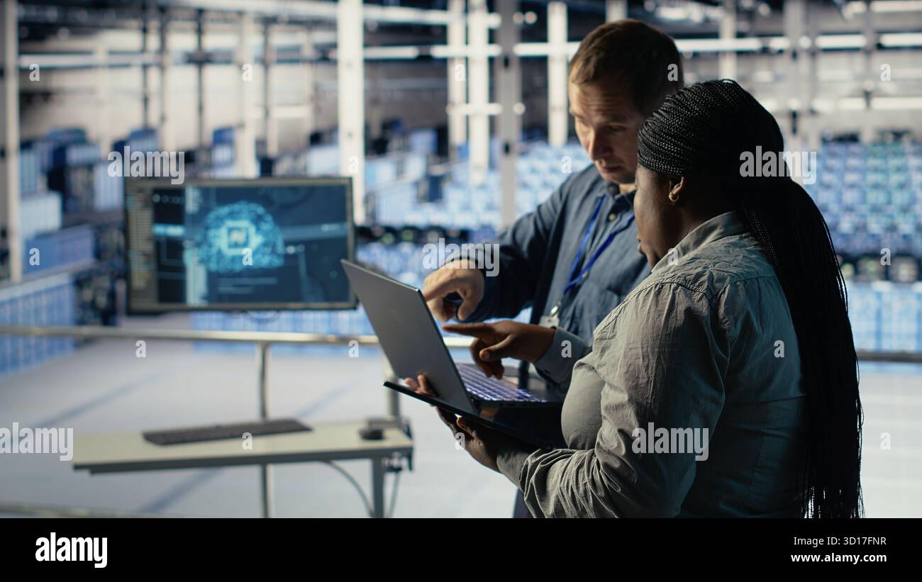 Server room coworkers use laptop to install updates and keep AI systems running. Data center engineering team uses notebook to write code improving infrastructure output, camera B Stock Photo
