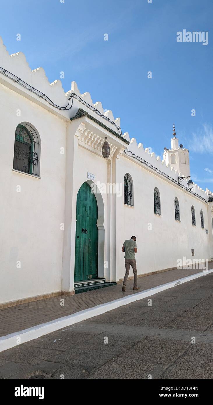 traditional white mosque exterior with serrated parapet, arched green door,and tall minaret under a blue sky, with a man walking past the building - Smartphone Captured Stock Image