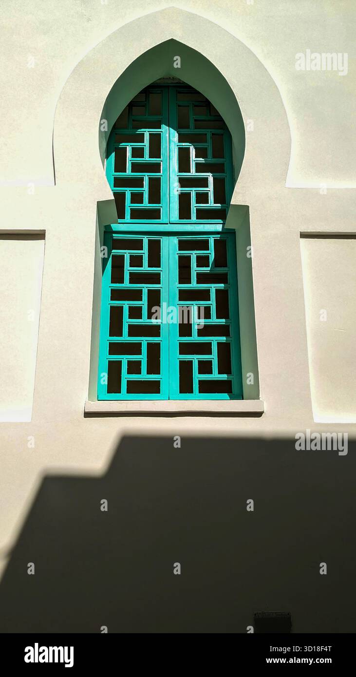 Traditional arched window with geometric turquoise lattice casting shadows on a white stucco wall under bright sunlight - Smartphone Captured Stock Image