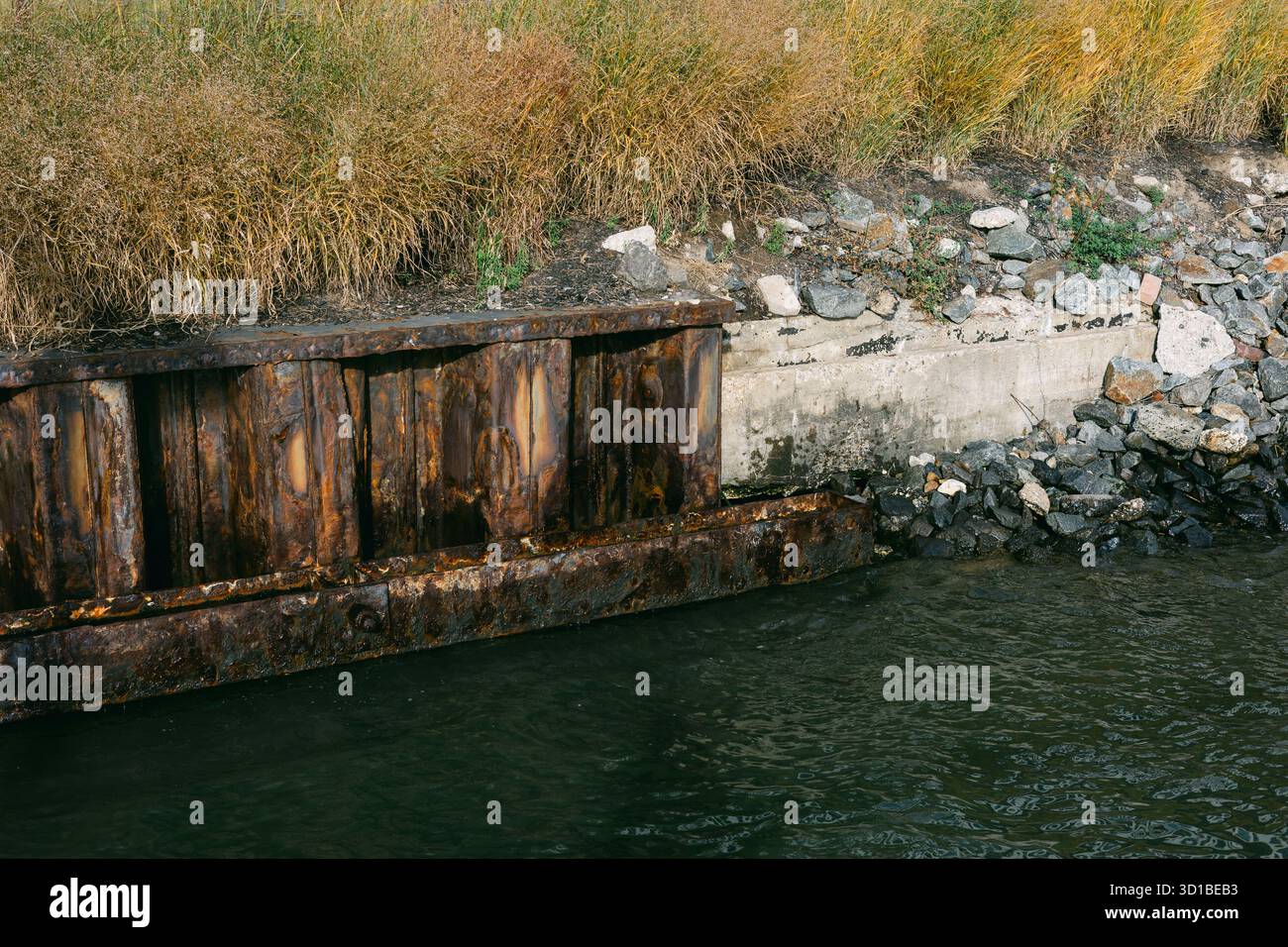 Rusty dock structure meets calm water, framed by lush grass and stones on a sunny day, creating a tranquil scene. Stock Photo
