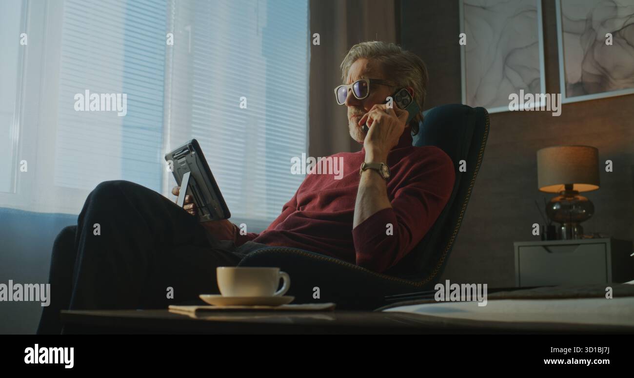 Gray Haired Businessman Sits in Modern Hotel Room With Tablet On Lap, Talking on Phone as Breakfast Served. Concept of Remote Work, Business Travel, and Digital Communication on Go. Handheld. Stock Photo