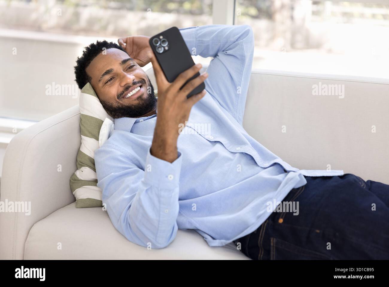 Relaxed African guy using smartphone while lying on cozy sofa Stock Photo