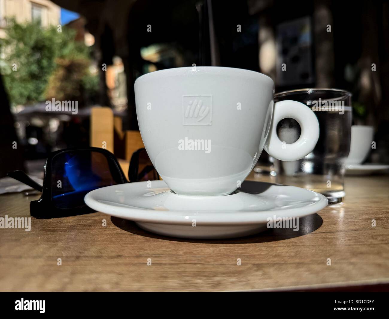 Close-up of an Illy espresso cup on a wooden outdoor café table with sunglasses and a glass of water - Smartphone Captured Stock Image