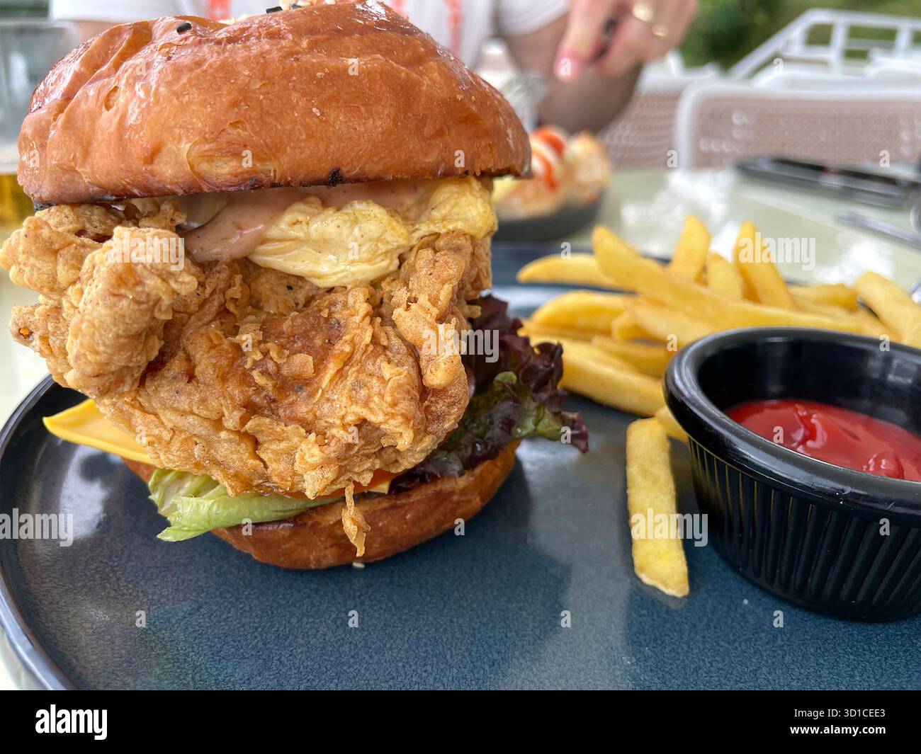 Close-up of a crispy fried chicken sandwich with lettuce, cheese, and sauce served with French fries and ketchup on a dark blue ceramic plate - Smartphone Captured Stock Image