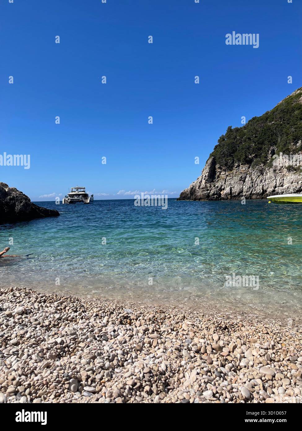 Idyllic Secluded Pebble Beach Cove with Crystal-Clear Turquoise Water and Rocky, Tree-Covered Cliffs Under a Deep Blue Summer Sky, with a Tourist Boat - Smartphone Captured Stock Image