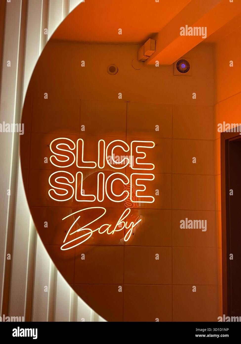 Warm Orange Neon Sign Reading 'SLICE SLICE Baby' on a Tiled Wall, Reflecting a Circular Light Fixture in a Trendy Pizzeria or Restaurant - Smartphone Captured Stock Image