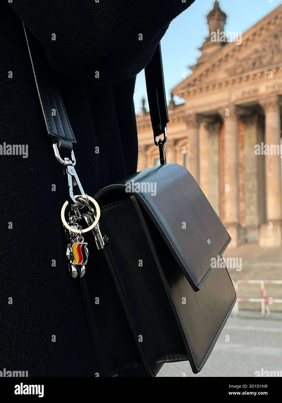Close-up of a person wearing a black coat and a black leather shoulder bag with a German flag keychain, standing near the historic Reichstag building - Smartphone Captured Stock Image