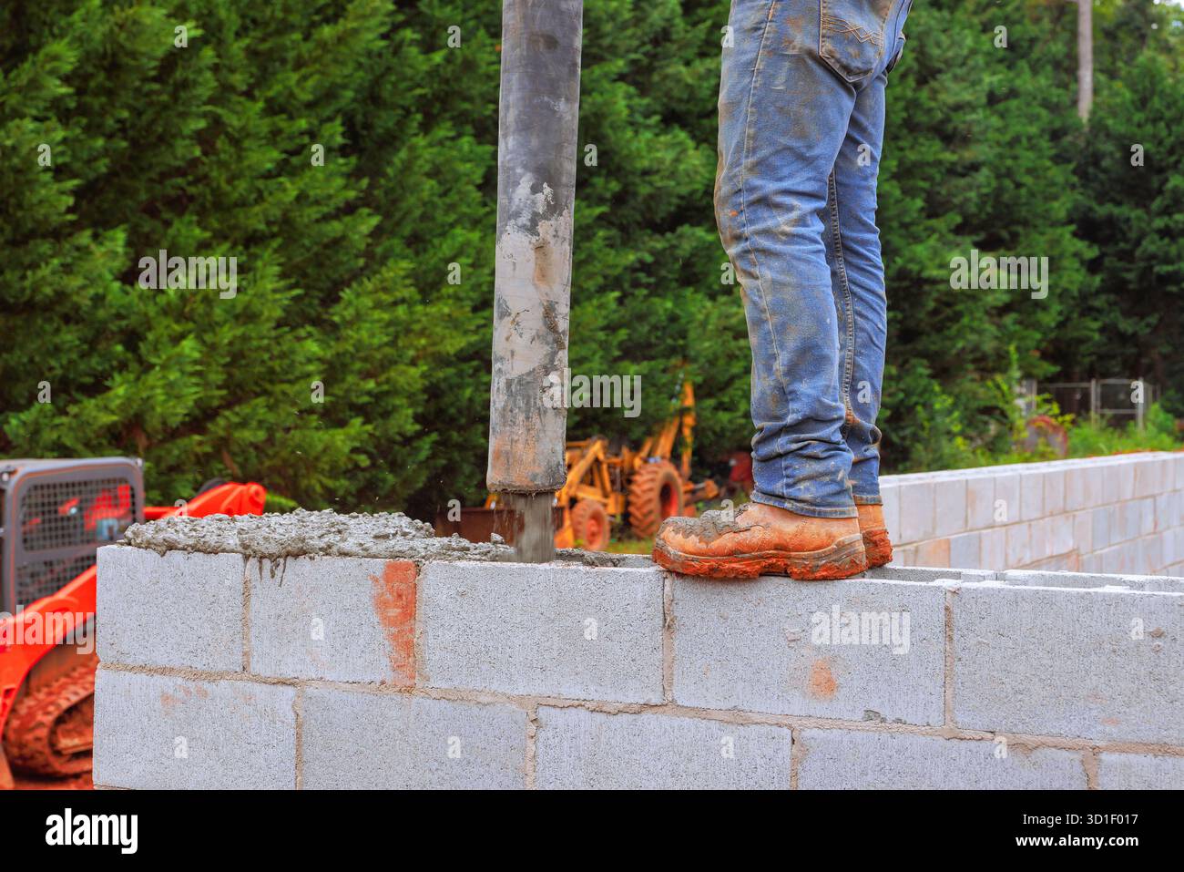 Worker stands on wall, directing concrete into block structure using pipe while surrounded by equipment. Stock Photo