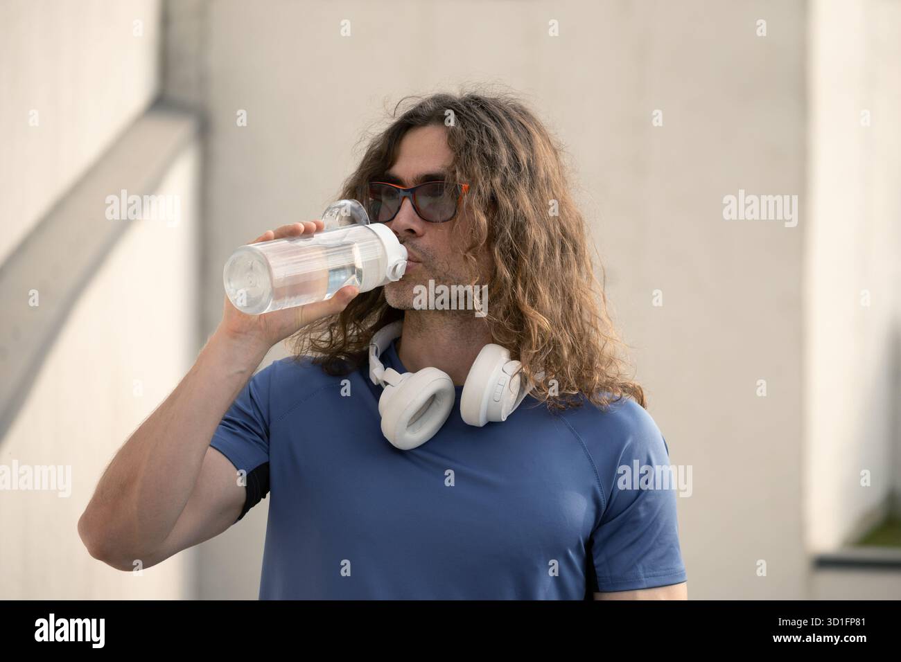 Handsome sporty fit athlete drinking water from bottle in sunny day against urban white wall. Water balance. Stock Photo
