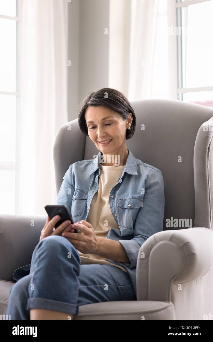 Smiling mature female using smartphone lounging in cozy chair Stock Photo