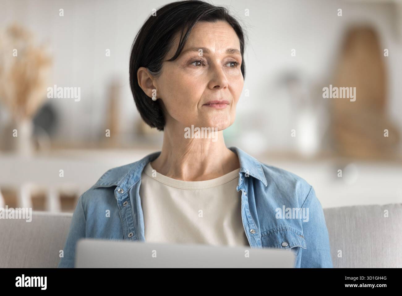 Thoughtful aged woman sitting on sofa looking above notebook screen Stock Photo