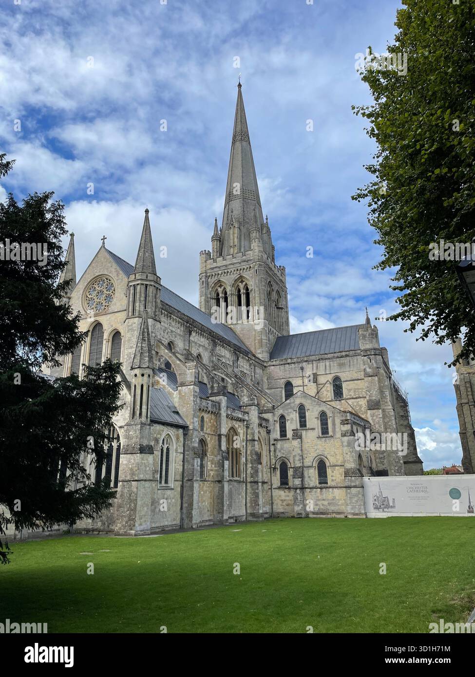 Picture of Chichester Cathedral between two trees with a grassed area in front and its spire pointing to a sky with wispy clouds. - Smartphone Captured Stock Image