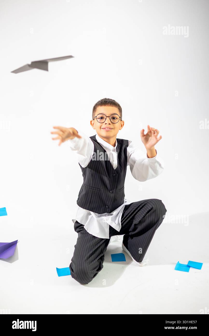 A joyful boy of Middle Eastern descent playfully throws a paper airplane in a studio, dressed in a striped suit and glasses. Stock Photo