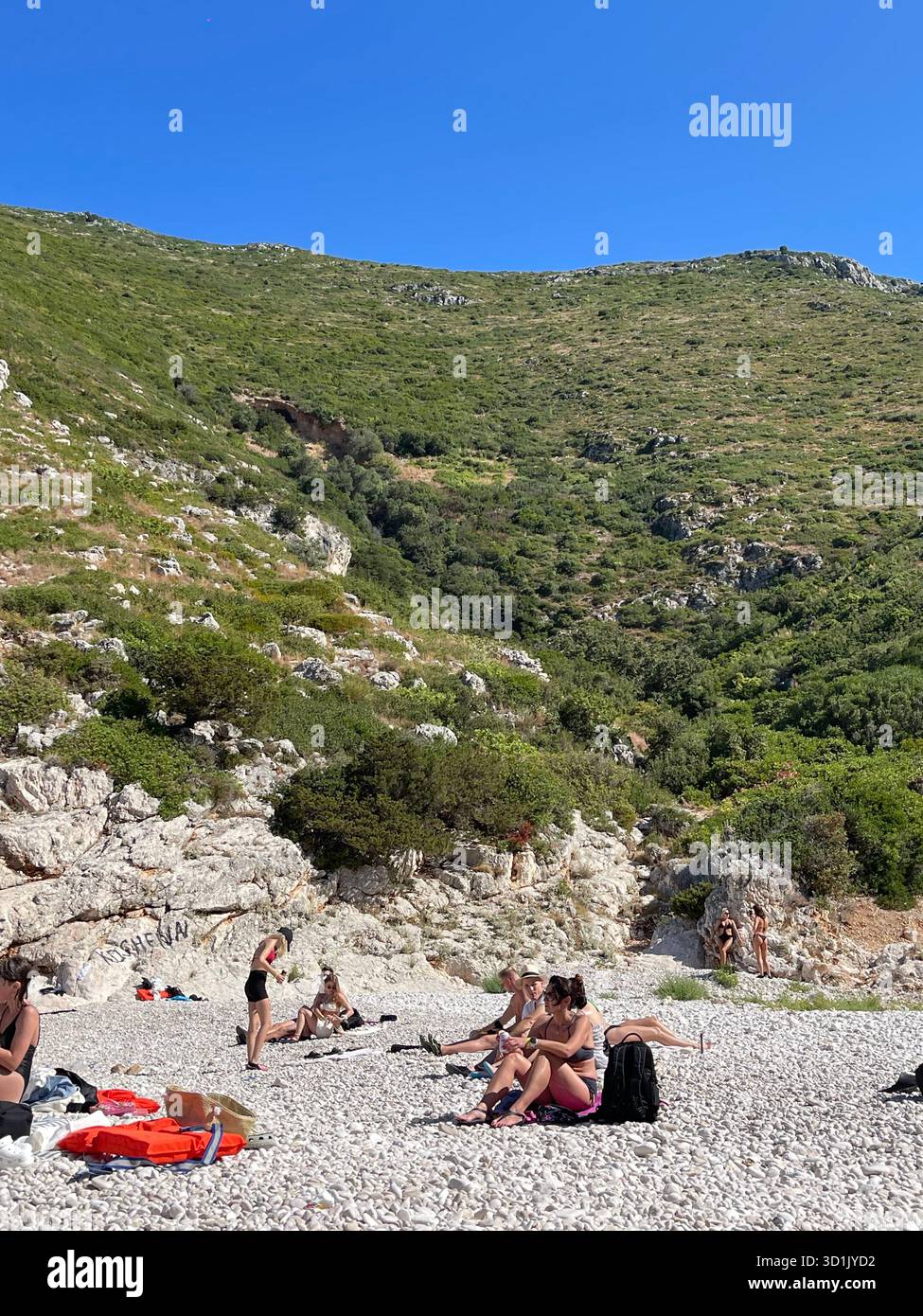 People relaxing on a pebble beach next to clear turquoise water, surrounded by massive rocks and green hills. Sunny day on the Mediterranean coast. - Smartphone Captured Stock Image