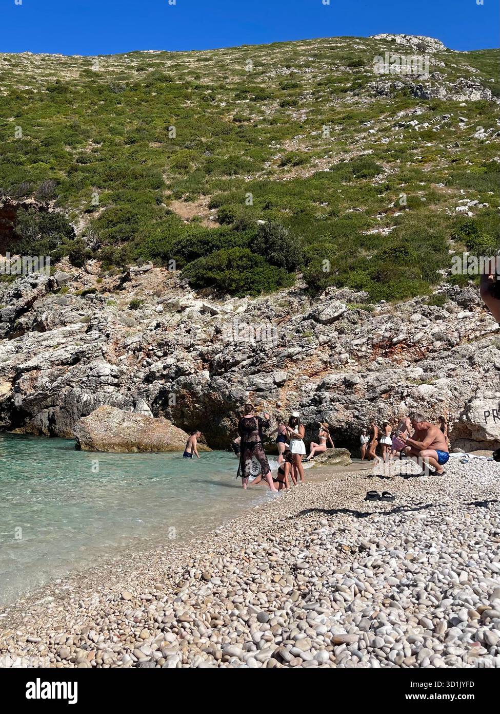 People relaxing on a pebble beach next to clear turquoise water, surrounded by massive rocks and green hills. Sunny day on the Mediterranean coast. - Smartphone Captured Stock Image