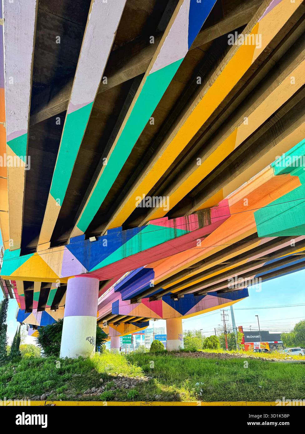 Colorful geometric mural painted on the underside of a highway overpass with greenery and urban surroundings below - Smartphone Captured Stock Image