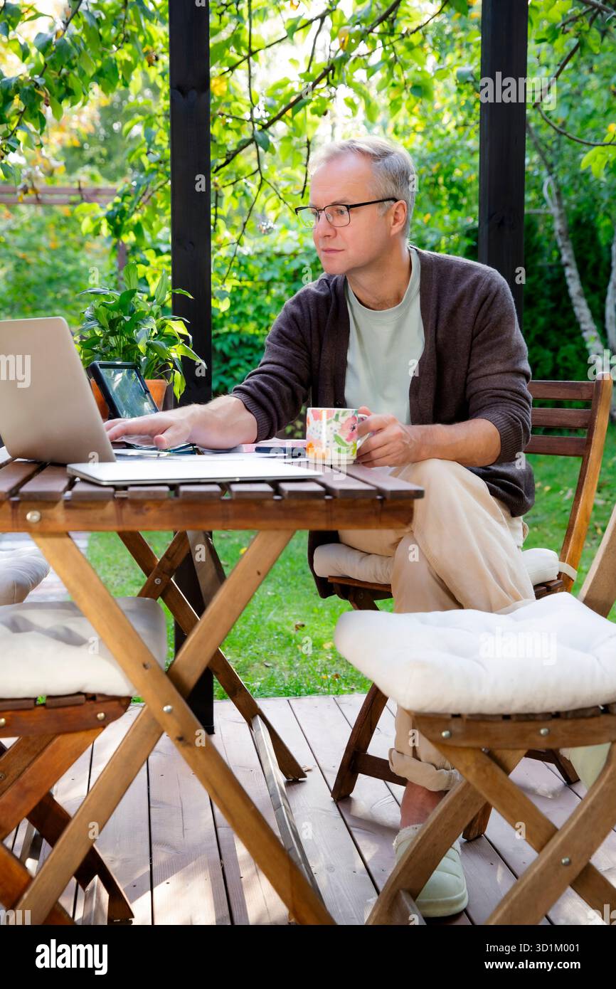 Middle aged man working remotely on laptop at cozy sunny patio, digital nomad lifestyle, outdoor home office, freelance and productivity concept Stock Photo