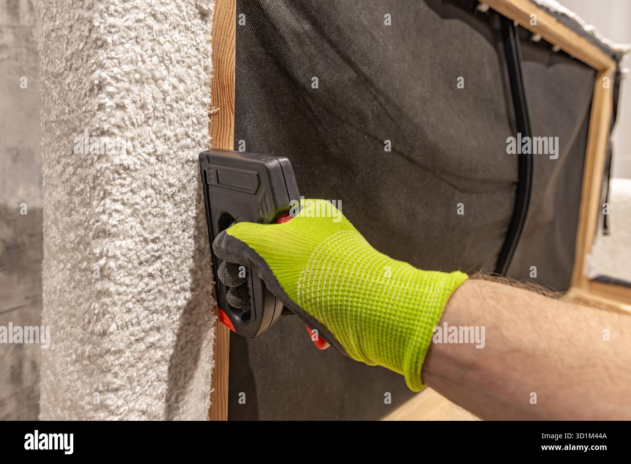 worker’s hand in green protective gloves using a black staple gun to attach light fabric to a wooden frame. Upholstery repair or furniture renovation Stock Photo