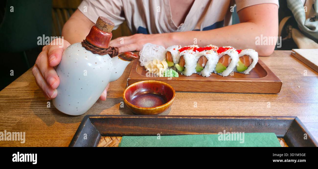 Hand holding a traditional sake bottle next to a beautifully arranged sushi platter with wasabi and pickled ginger, showcasing culinary art. - Smartphone Captured Stock Image