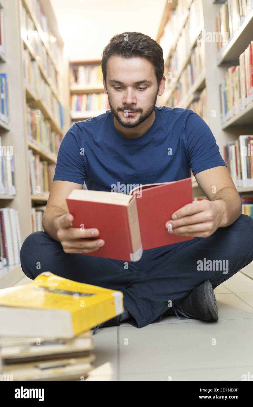 Handsome young student sitting on library floor reading book in college Stock Photo