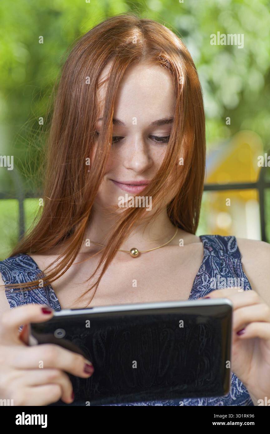 Redhead student leaning against a tree using her tablet on college campus. Redhead woman with tablet outdoors Stock Photo