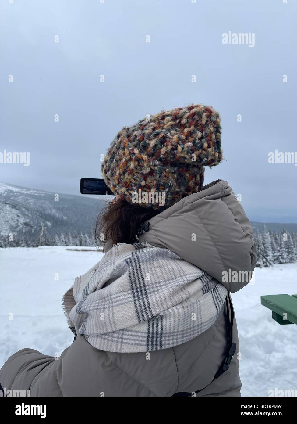 Person wearing a warm winter puffer jacket, plaid scarf, and a knitted bobble hat, enjoying a snowy mountain landscape. View from behind. Winter touri - Smartphone Captured Stock Image