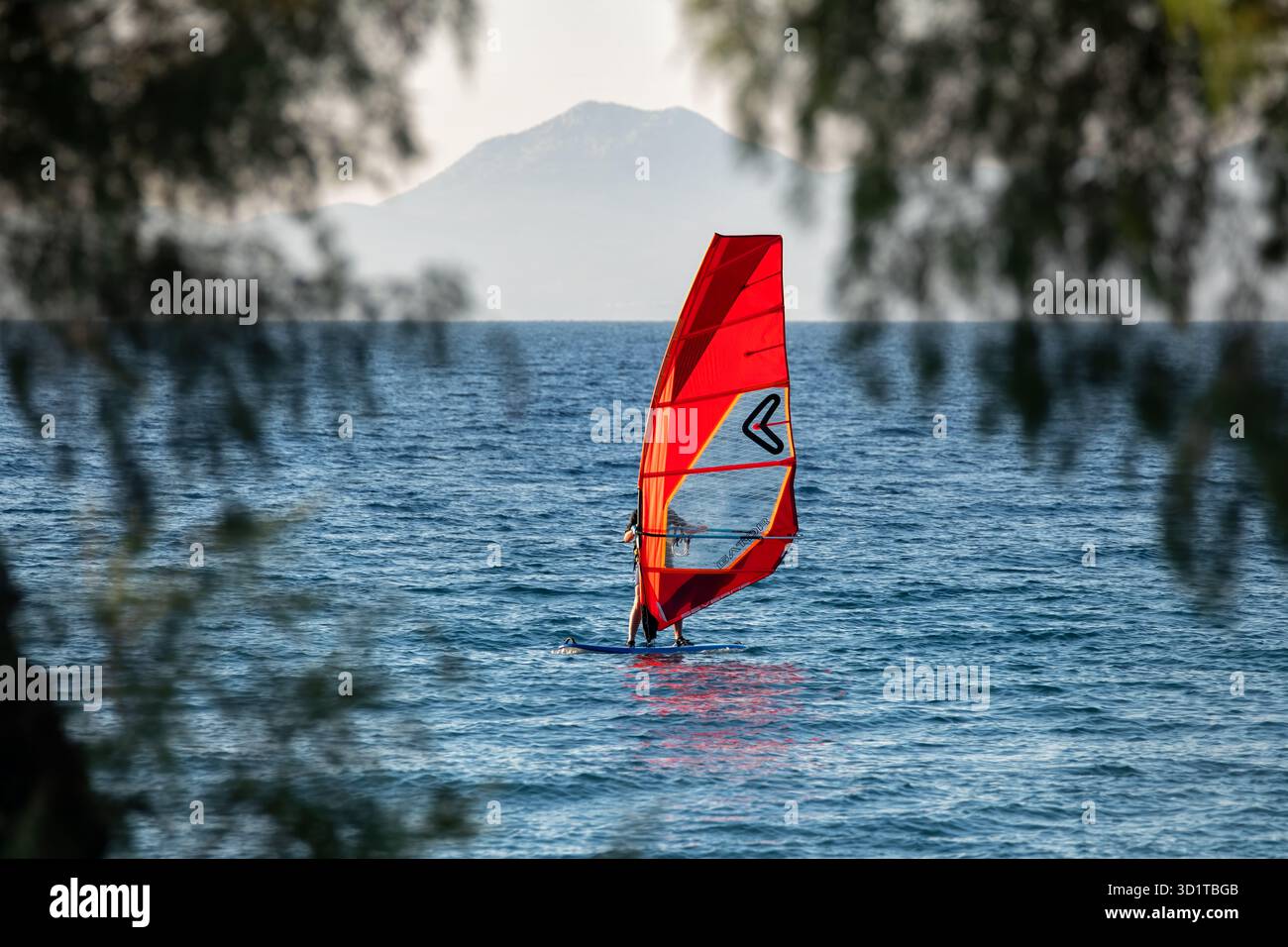 Rhodes, Greece: A wind surfer with a red coloured sail wind surfing close to the beach and shoreline. He's framed by coastal trees Stock Photo