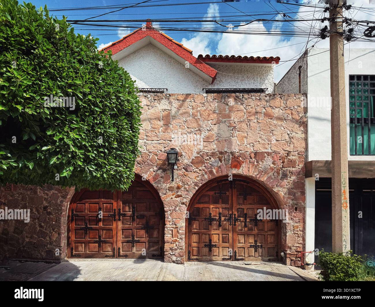 Stone facade house in Mexico with arched wooden garage doors and red-tiled roof in a residential street under sunny weather - Smartphone Captured Stock Image