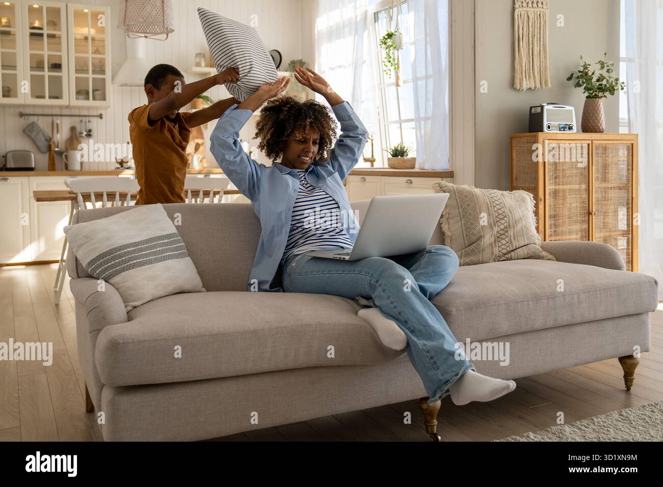 Angry mom brushes off with hand son play with pillow distract from remote work on couch with laptop Stock Photo