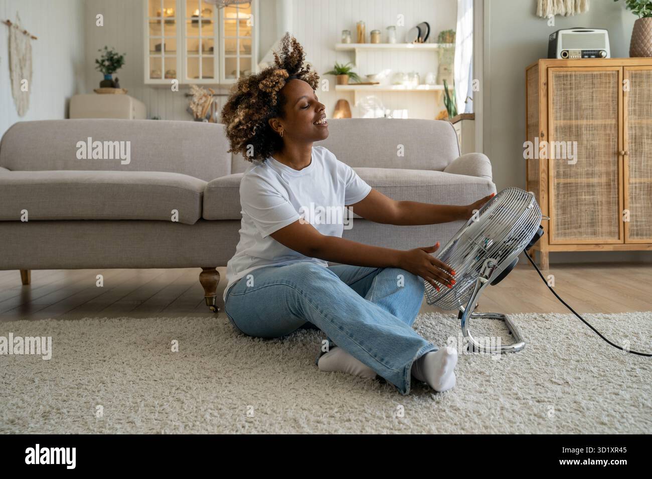Satisfied happy young African American woman using fan at home during extreme summer heat Stock Photo