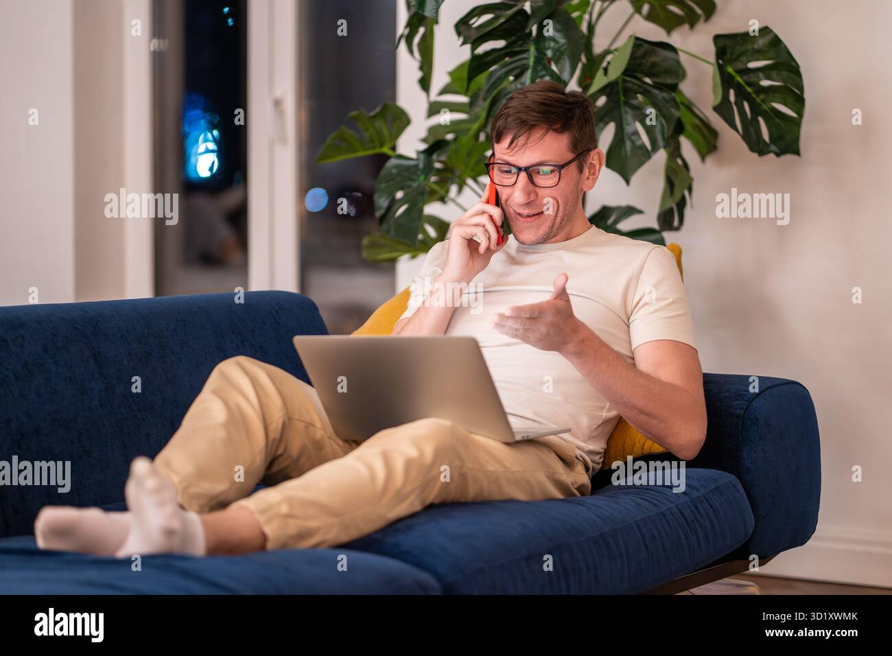 Smiling positive man freelancer talks phone works on laptop sits on couch at home in evening. Stock Photo