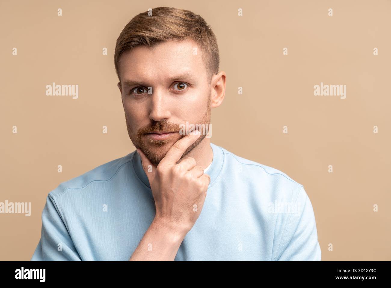 Thoughtful serious middle-aged bearded man touching chin and looking at camera on beige background Stock Photo