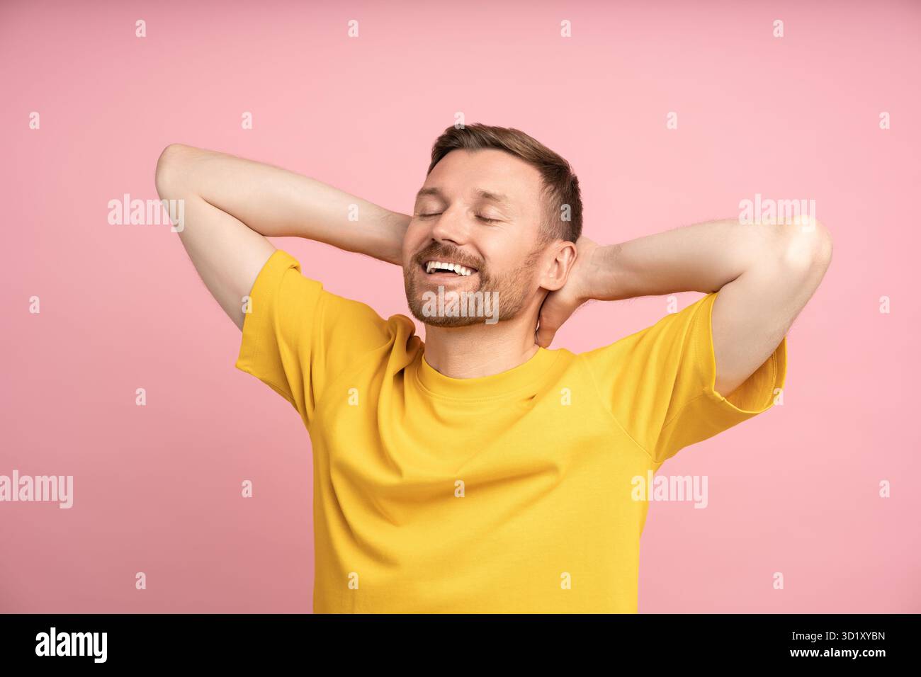 Relaxed man holding hands behind head smiling with closed eyes standing on studio pink background. Stock Photo