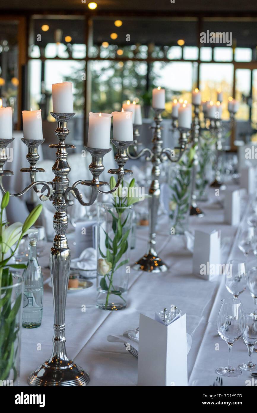 A beautiful dining arrangement features silver candelabras with lit candles and fresh flowers on tables set for an evening event in a well-lit venue w Stock Photo