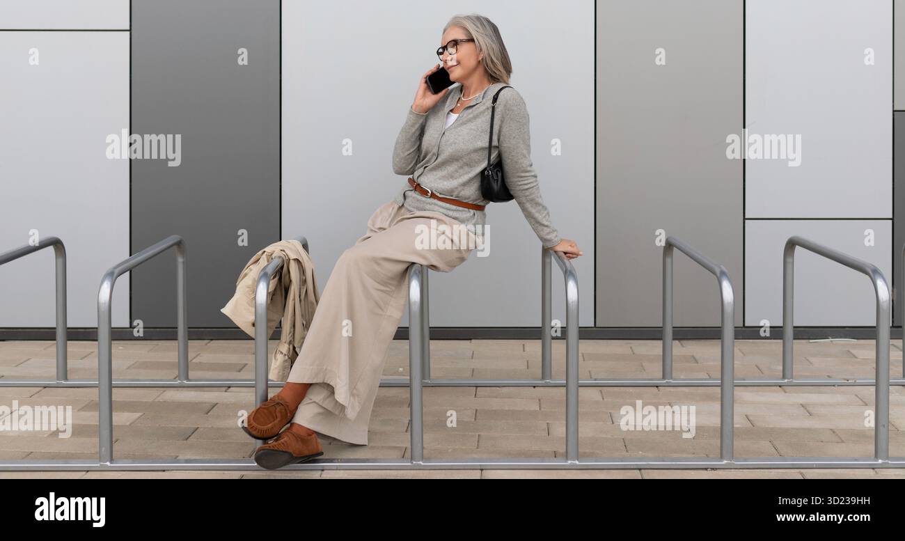 Older woman casually talking on mobile phone while sitting on railings outside modern building Stock Photo