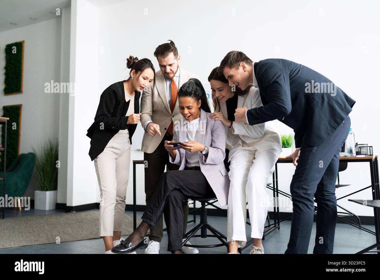 Employees in the office stand around a woman playing on a smartphone Stock Photo