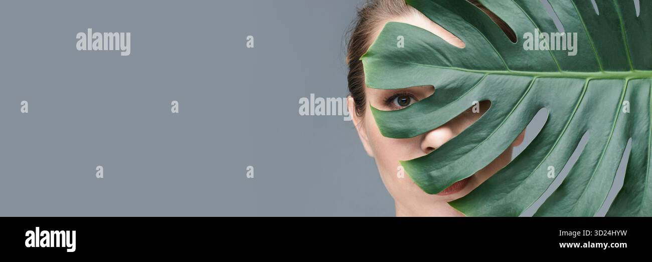 Caucasian female eye peeking through monstera leaf on grey background showing Stock Photo