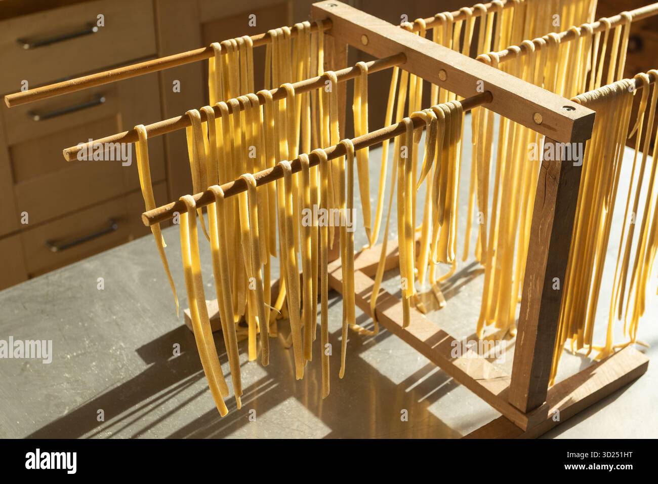 Homemade fresh Linguine pasta hanging out to dry, on a wooden rack, and backlit by sunlight. Traditional home cooking and healthy eating concept. - Stock Image