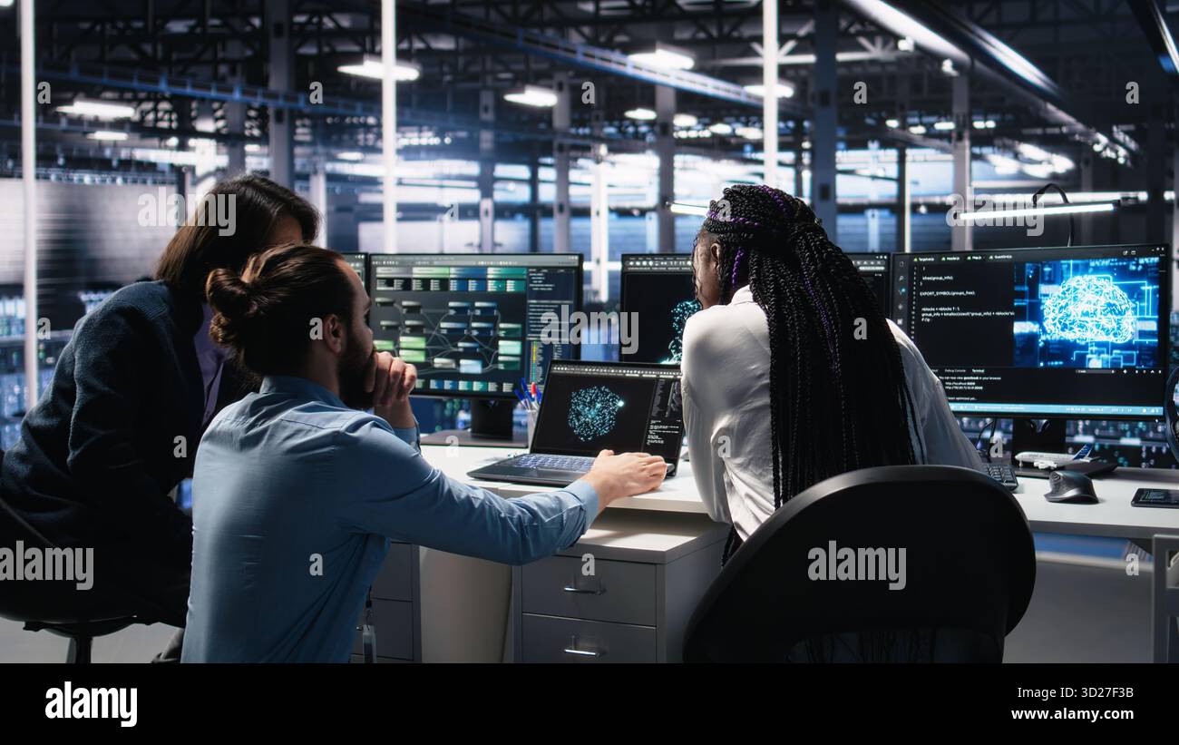 Teamworking data center technicians using machine learning algorithms to analyze large datasets. Server room colleagues gathered together at desk, collaborating, using AI neural networks, camera A Stock Photo
