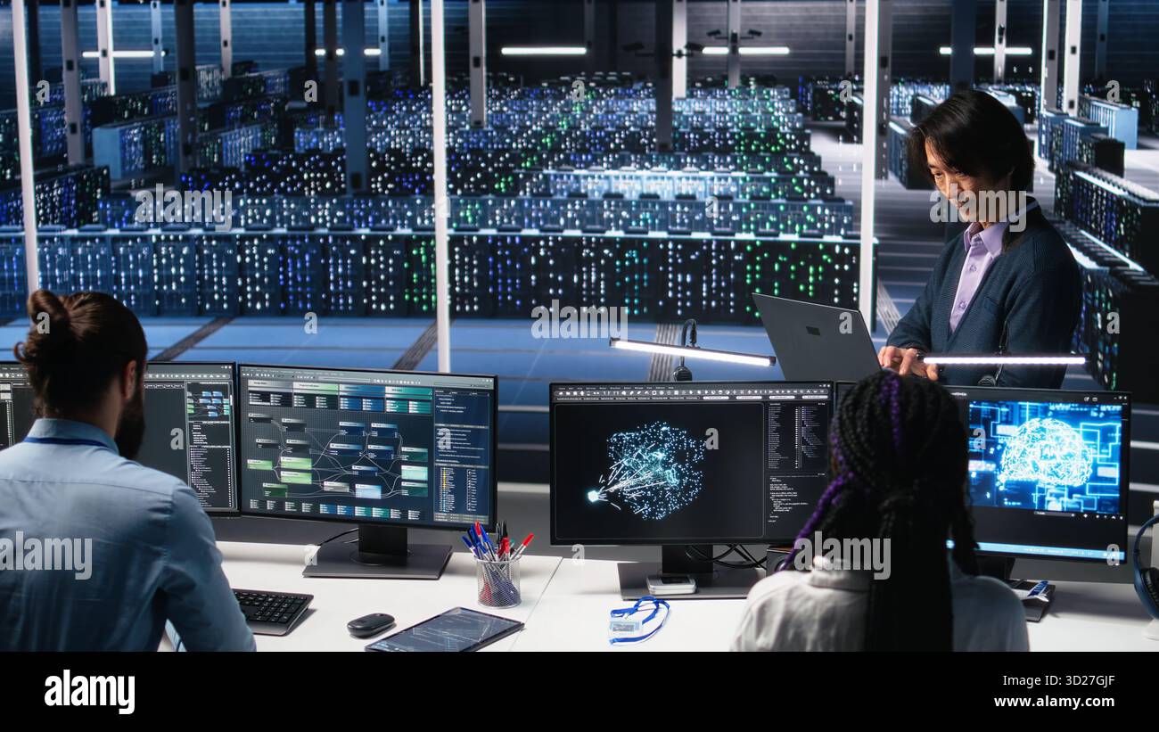Software developers cooperating in data center, reviewing AI diagnostics on computer. IT staff members discussing in server room, optimizing machine learning systems, camera A Stock Photo